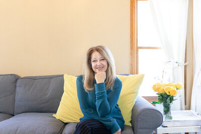 Cyndi Stewart sits smiling on a cozy gray couch with yellow pillows, beside a bright window and a vase of yellow roses.