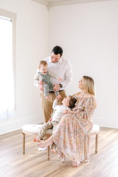 A family of four joyfully pose on a bench inside of a natural light studio while the two children tickle each other.