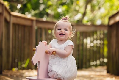 A cheerful baby girl in a white dress sits on a pink wooden horse, embodying the essence of childhood joy.