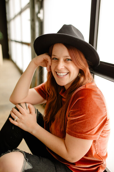 Photo of a red-haired woman sitting smiling, wearing a black hat with an orange T-shirt and black pants. Her hand is on her cheeks