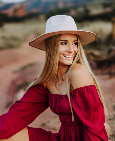 A girl in a red dress and a hat looks over her shoulder while posing for senior photos at Red Rocks.
