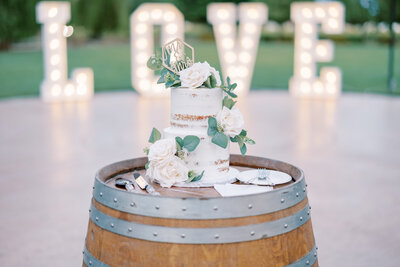 bay area photographers photographs white nake wedding cake with large white roses and greenery sitting on a barrel at a wedding reception in Sonoma