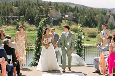 Bride and groom walking up the aisle during an outdoor Colorado mountain wedding as guests toss white confetti, with pine trees and rocky cliffs in the background. Ceremony at Perry Park Country Club