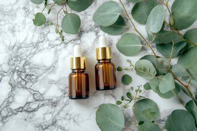 Two amber essential oil dropper bottles with gold caps surrounded by eucalyptus leaves, reflecting the natural luxury of Ember to Sage's wellness experience