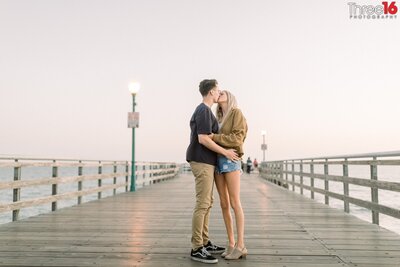 Engaged couple share a kiss while standing on the Seal Beach Pier