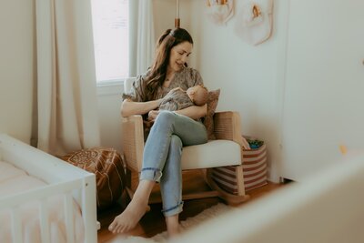 A woman sits in a beige chair, holding a newborn wrapped in a patterned blanket. She is in a softly lit room with curtains and a crib nearby.