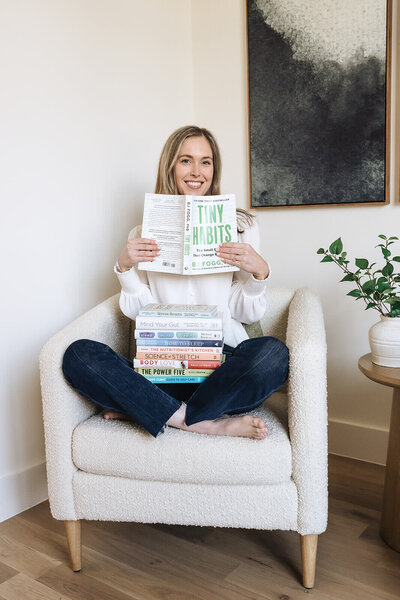 Wellness coach reading Tiny Habits book with a stack of health and nutrition books—supporting simple, sustainable habits for moms.