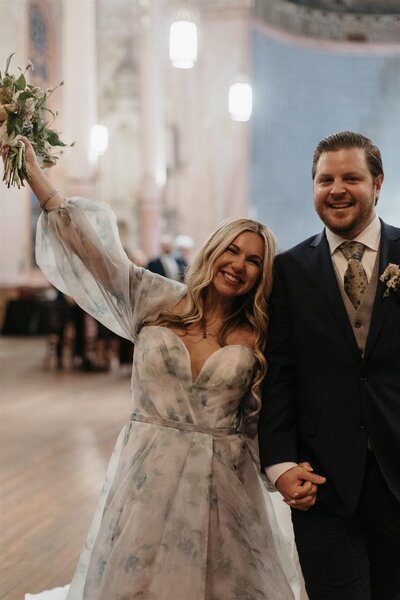 The same bride and groom walking down the aisle, the bride raising her bouquet and smiling widely.