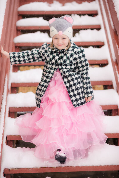 Little girl dressed in a pink Princess dress and  houndstooth jacket with mouse hat walking in the snow.  Taken by  Dripping Springs Texas based Lydia Teague Photography.