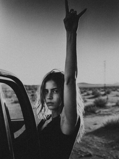 A person stands by a vehicle in a desert, raising one arm and making the "rock on" hand gesture. The black-and-white image captures the bold energy behind Sweet Pea Ink Creative’s brand design vision.