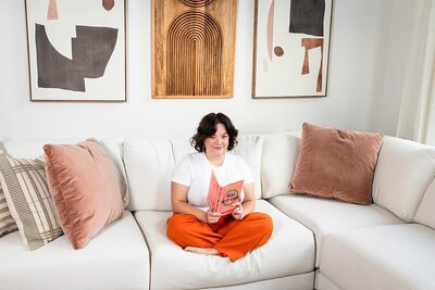 Woman in a white t-shirt and orange pants sits cross-legged on a cream-colored couch, reading Real Self-Care during a personal branding photoshoot. Captured in a bright, modern living space with abstract art and soft textures, this image conveys calm, confidence, and intentional lifestyle branding.