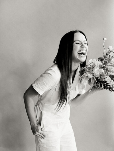Black and white photo of a woman in a jumpsuit holding flowers and laughing in a studio