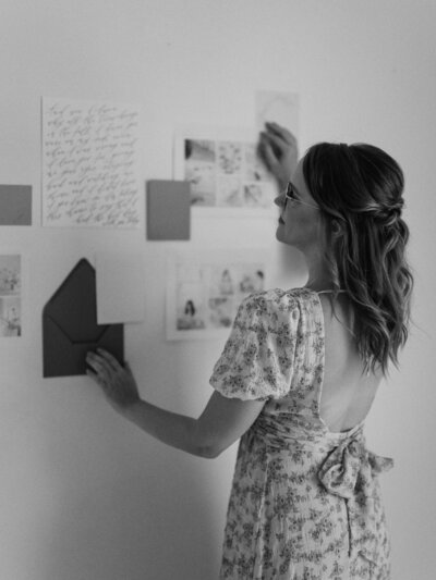 Black and white photo of Jen Krause arranging paper and stationery on a wall