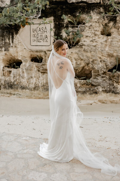 A bride in a backless white gown poses on a sandy beach with a long veil flowing behind her. She stands in front of a rocky backdrop with a ‘La Casa de la Playa’ sign visible on the wall.