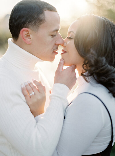 Up close photograph of a couple at their winter engagement session. They are standing nose to nose as if they are almost about to kiss. They are looking at each other with mouths parted just slightly. He is cupping her chin softly and she has her hand up on his chest. They are both wearing ivory and have black hair. The sun is peaking in just over the tops of their heads to give a romantic light and mood. Photographed by wedding photographers in Charleston  Amy Mulder Photography