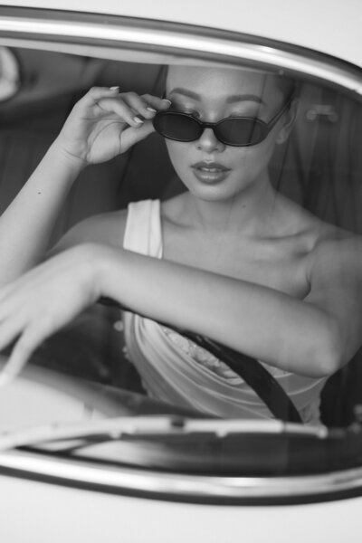 A woman with sunglasses is positioned in a car, embodying a chic and carefree attitude.