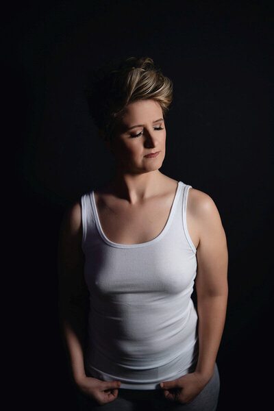 A woman with short hair stands against a dark background, wearing a white tank top. Her eyes are closed, expression peaceful, lighting soft and dramatic.