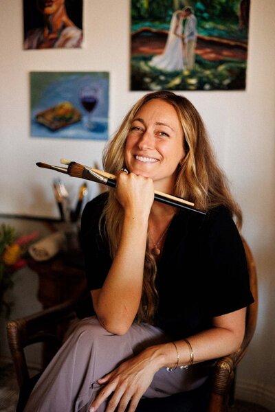 Artist smiles with a handful of paint brushes in her Maui studio