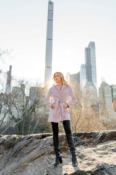 woman standing on a rock with her hands in her coat pockets with the city skyline behind her
