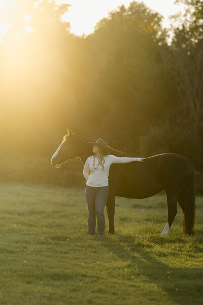 a girl and a black horse standing in a field at sunset looking off into the distance