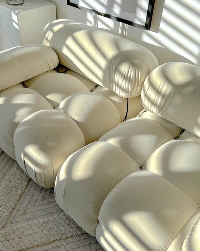 A close-up of a luxurious, cream-coloured modular sofa (reminiscent of a Mario Bellini Camaleonda) bathed in soft, dappled natural sunlight. The interplay of light and shadow highlights the plush, textured upholstery, illustrating how natural light influences the perception of neutral colours and enhances the atmospheric and emotional resonance within an interior space, aligning with principles of colour psychology for wellbeing.