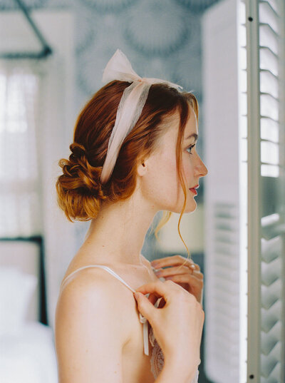 Bride looking in the mirror as she gets ready for her wedding wearing a gossamer silk pink bow around her hair