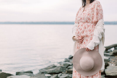 Pregnant mother wearing a floral dress and holding a hat while standing on a rocky beach