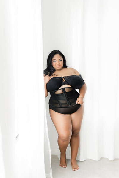 Curvy woman wearing off the shoulder black bodysuit standing posing in boudoir photoshoot