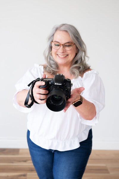 Woman photographer with grey mid length hair looking at back of camera, smiling.