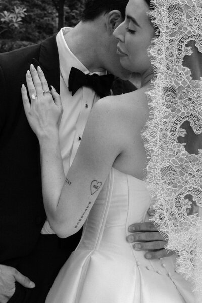 Black and white portrait of a bride and groom wrapped in a delicate lace veil. The groom leans in with a soft expression while the bride gazes forward, holding a bouquet. A moody, artistic documentary wedding moment 