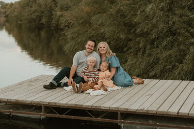 Photo taken by Bailee Freeman Photography of a family with a mom, dad, son, and daughter. The family is sitting on a boat dock in rexburg, Idaho and snuggled up to each other and smiling at the camera. 