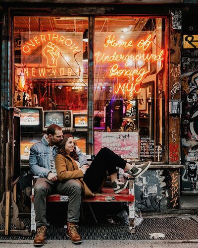 Couple snuggle together on a bench outside a store in a busy city
