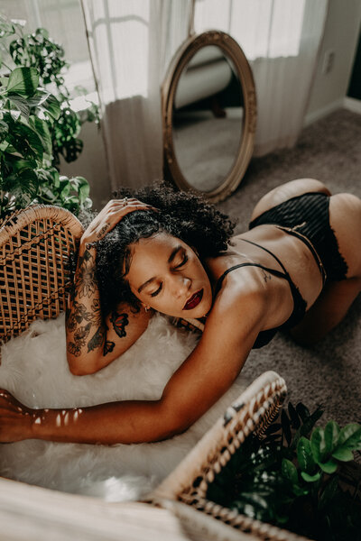 Black Mexican woman in black lingerie, leaning on chair with sheepskin, confident and sensual boudoir portrait.