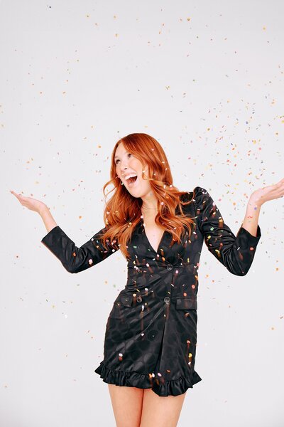 Redheaded woman in a chic black wrap dress throwing colorful confetti in the air during a playful studio branding session.