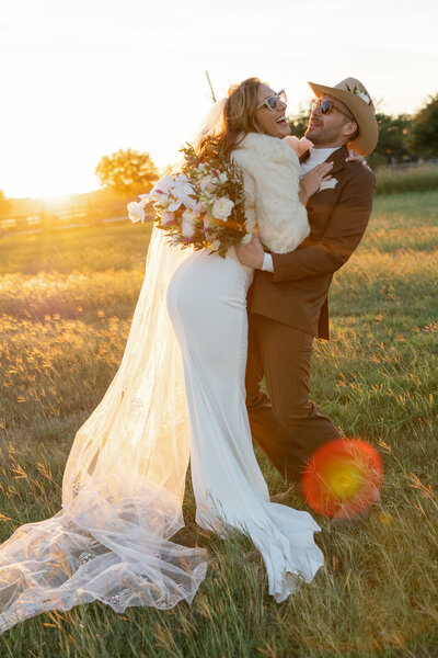 Bride being lifted up by her groom laughing and throwing her head back wearing sunglasses and a fur coat while her veil trails along catching all the sun light.