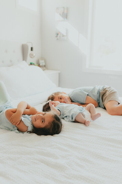 Photo of 2 older siblings laying on the bed with their baby sister in between them.