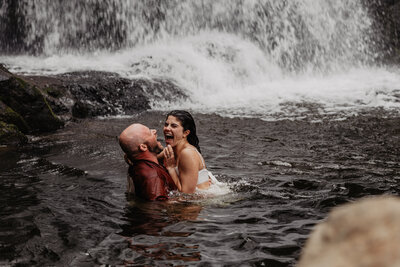Bride and groom playing in water below waterfall