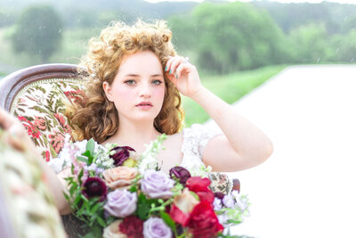 bride with flowing curls holding a lush bouquet of red and purple flowers, seated outdoors at Willow and Stone Events.