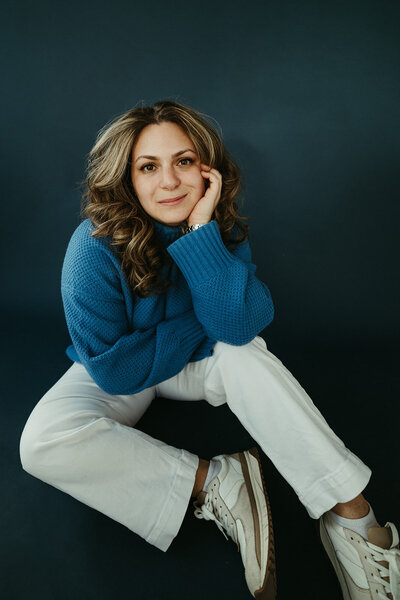 Sara Dobbins sits on blue photo backdrop leaning forward onto her hand and knee looking softly at camera