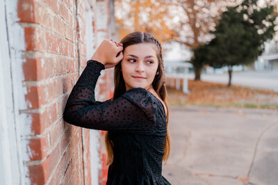 A high school senior girl poses for the camera leaning against a brick wall and smiling off into the distance.