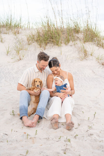 couple sits on the beach in front of the dunes, father holds family dog while mother holds baby wearing a white bow on topsail island, north carolina