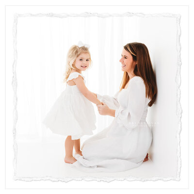 The photo shows a mom and daughter during a Motherhood portrait photo session. They are both dressed in all-white outfits and standing in an all-white studio. The mother and daughter are holding hands and smiling, creating a heartwarming image. The photo was taken in a clean, pure style by photographer Bri Sullivan.