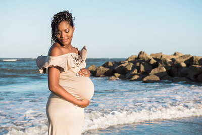 Expecting mother in a neutral off-shoulder gown stands at the shoreline, bathed in golden hour light with ocean waves and rock formations behind her.
