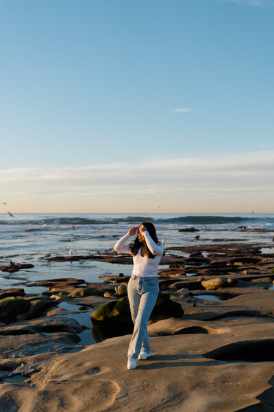 Lauren Ahn, photographer, standing on the rocky shoreline of a beach in San Diego, shielding her eyes from the sun while looking out over the ocean during golden hour.