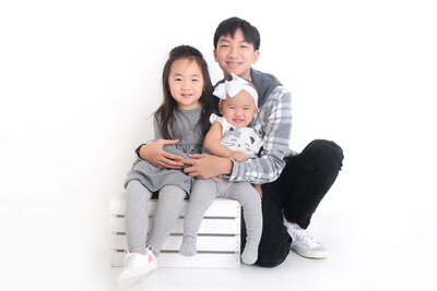 a baby girl and her two siblings cuddle up and smile during their family photoshoot