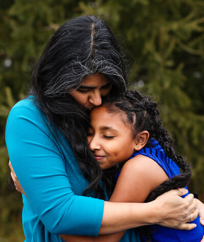 A women in blue hugs and kisses a young girl, both standing in front of trees during a portrait session in Kingston.