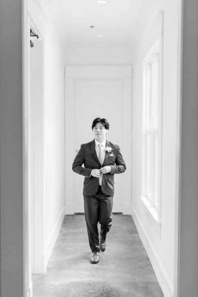 A black and white image of a groom walking down a white hallway buttoning up his jacket before he see his bride for the first time