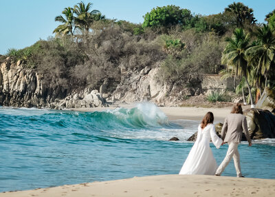 A bride in a flowy silky wedding dress and a groom in tan light suit walk away from the luxury elopement photographer looking at a huge, beautiful blue wave curling and about to crash ashore during their Mexico beach elopement.  There are rocky cliffs and palm trees visible on the beach across from where they are walking. 