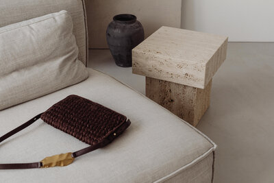 A chic, modern interior featuring a beige sofa with a stylish black leather shoulder bag placed on top. A marble side table sits beside it, accompanied by a tall grey vase on the floor. Designed for a home decor brand, this image captures a neutral, sophisticated aesthetic perfect for social media branding.