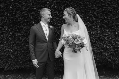 Black and white image of bride and groom in front of  greenery holding hands and smiling at each other